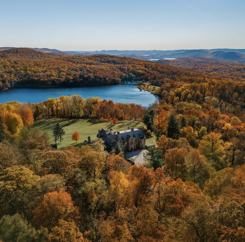 Aerial view of a stone castle amid vibrant autumnal trees, with a blue lake and rolling hills in the distance under a clear sky.