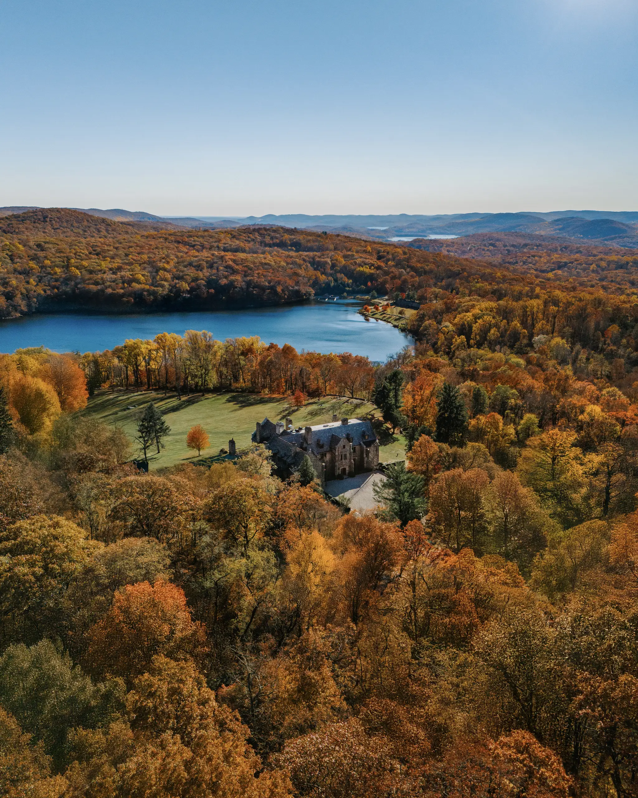 Aerial view of a stone castle amid vibrant autumnal trees, with a blue lake and rolling hills in the distance under a clear sky.