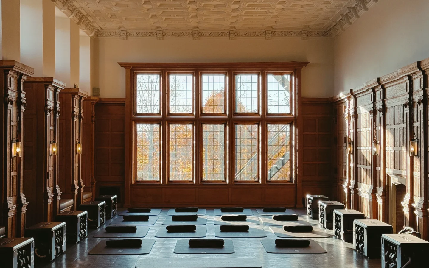 Yoga class setup in a grand room with ornate coffered ceiling, wooden paneled walls, and large windowed back wall; mats and bolsters arranged for practice.