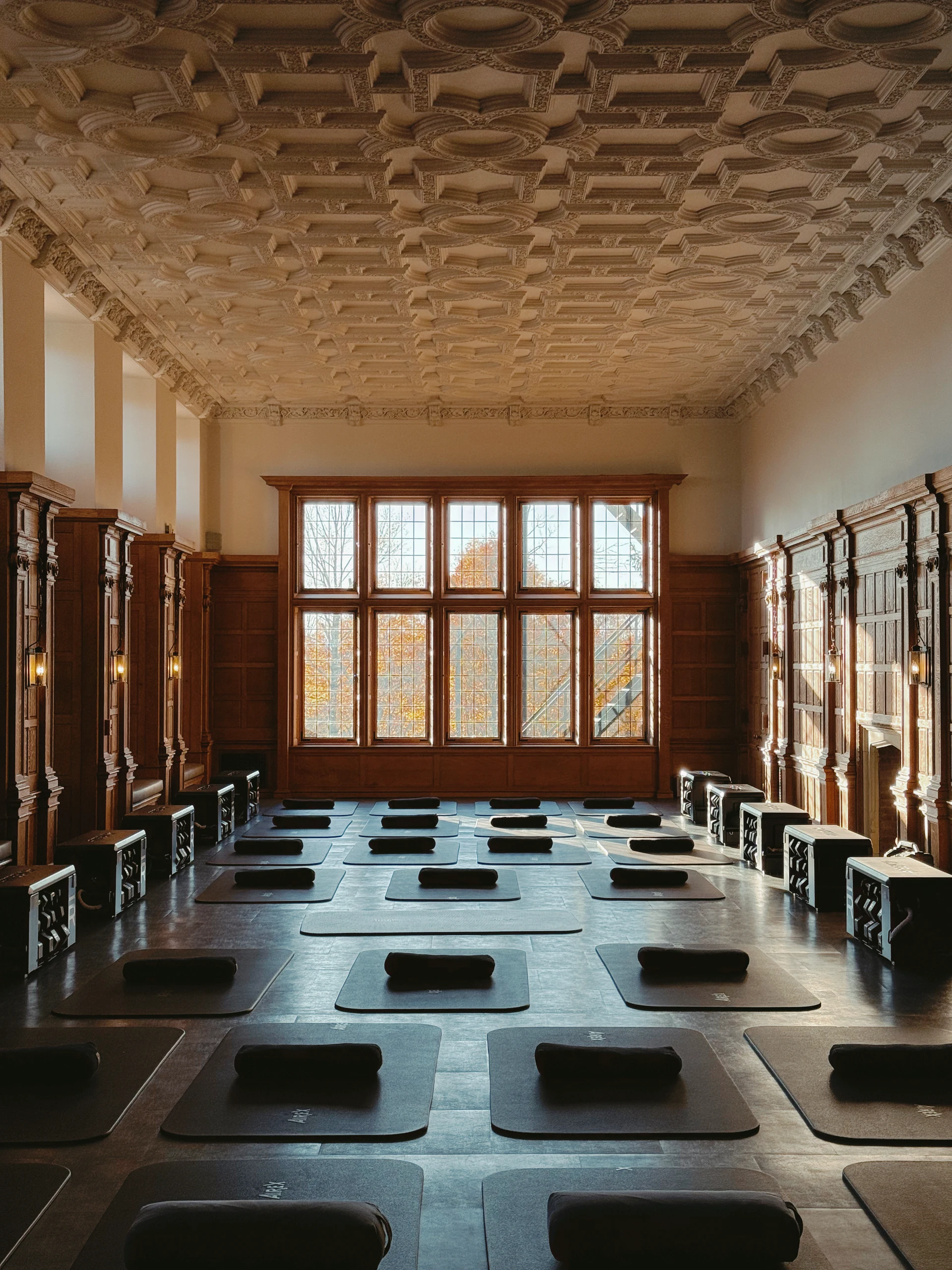 Yoga class setup in a grand room with ornate coffered ceiling, wooden paneled walls, and large windowed back wall; mats and bolsters arranged for practice.