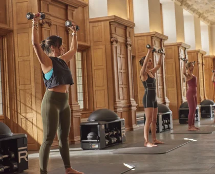 Group fitness class doing overhead dumbbell presses in a wooden-paneled studio; participants stand on mats and lift dumbbells above their heads.
