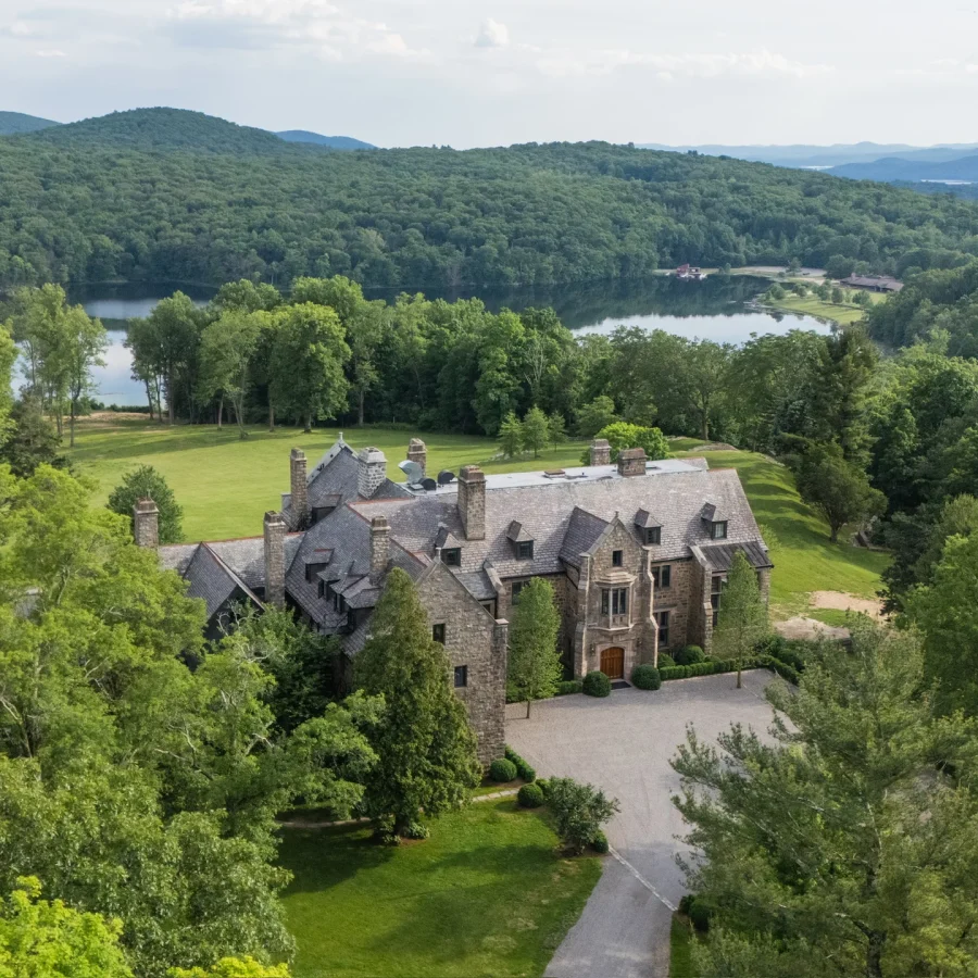 A large stone estate with a castle-like roof amid dense trees, with a lake and forested hills in the distance.