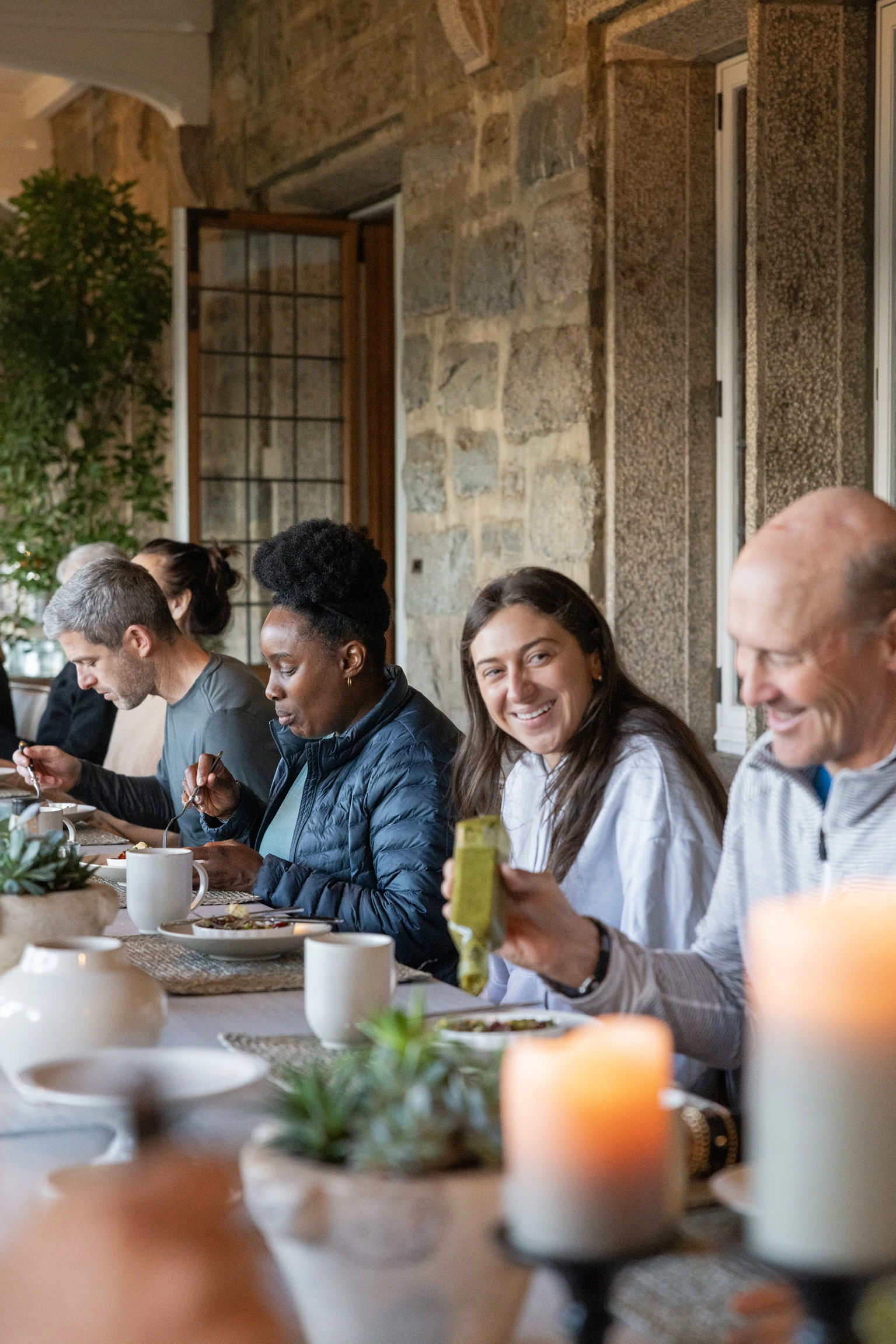 Group of people dining at a long table in a stone-walled dining room, smiling and eating together.