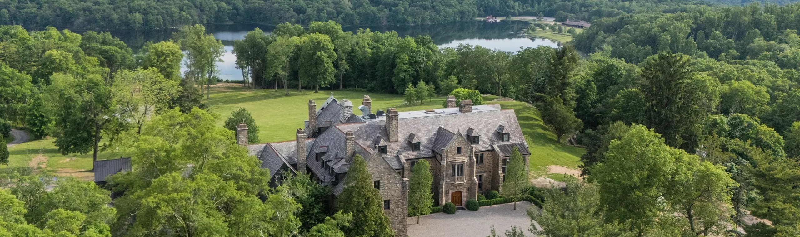 A stone manor with turrets sits among green trees, with a lawn and gravel drive in front and a forested lake beyond.