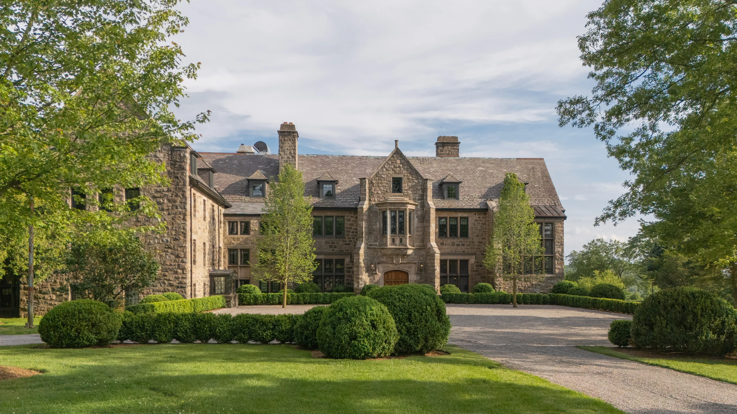 Stone manor with symmetrical facade, manicured hedges, and gravel driveway framed by green trees under a blue sky.