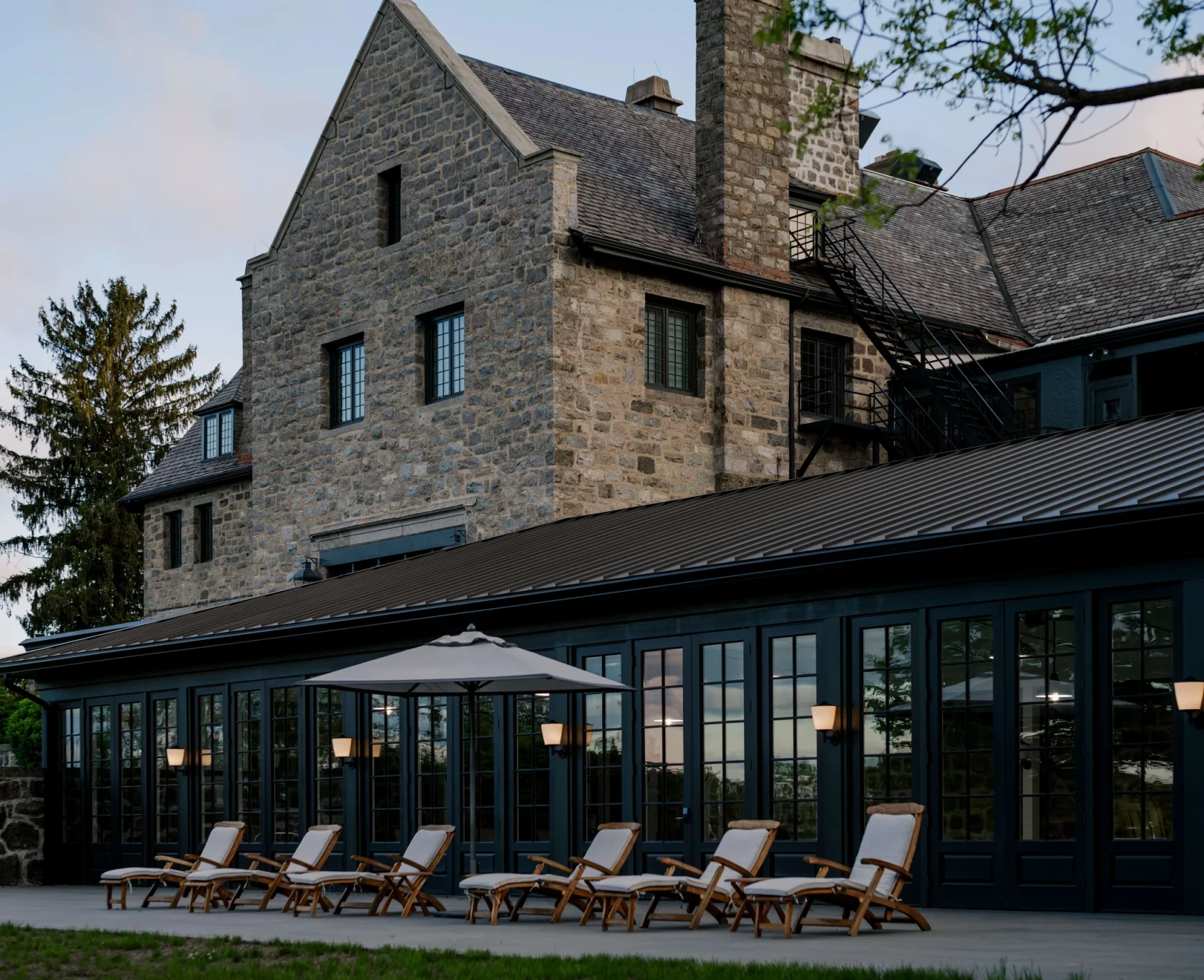 Stone building with brick-and-glass conservatory and row of wooden lounge chairs on a patio outside at dusk.
