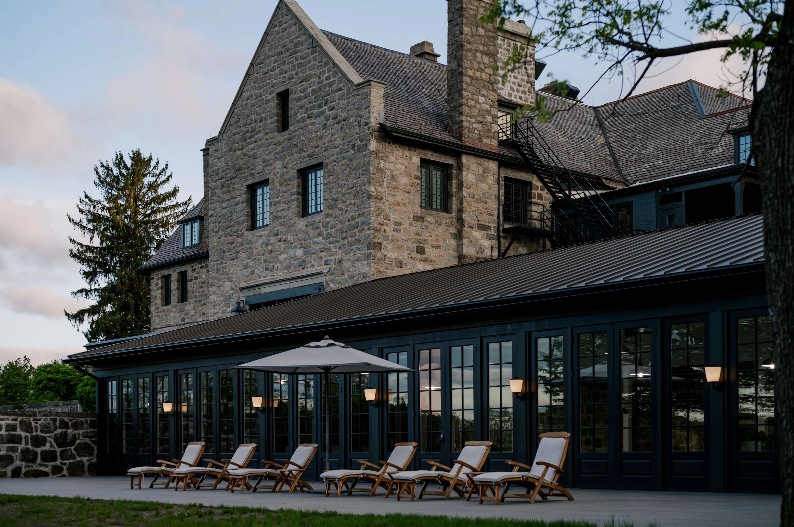 Stone building with brick-and-glass conservatory and row of wooden lounge chairs on a patio outside at dusk.