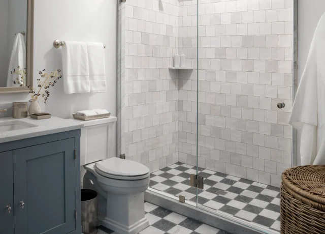 Modern bathroom with a blue vanity, white toilet, and a glass-enclosed shower featuring white subway tiles and a black-and-white checkered floor.