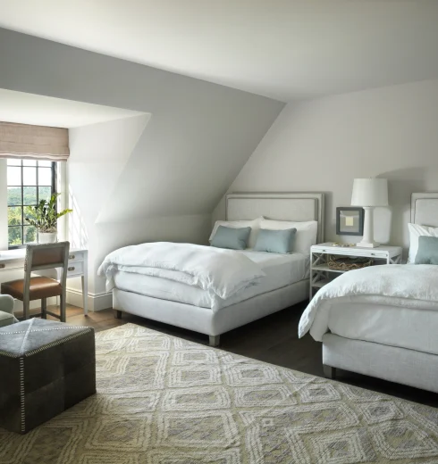 Modern bedroom with two white upholstered beds, matching nightstands, and soft gray walls by a large window with beige shade.