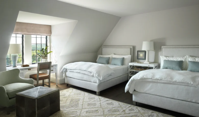 Modern bedroom with two white upholstered beds, matching nightstands, and soft gray walls by a large window with beige shade.