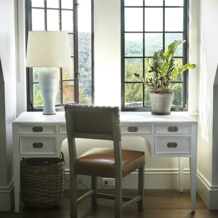 White write desk with a gray upholstered chair, a white ceramic lamp on the left, and a potted plant on the right by a large grid-window showing a hilly landscape.