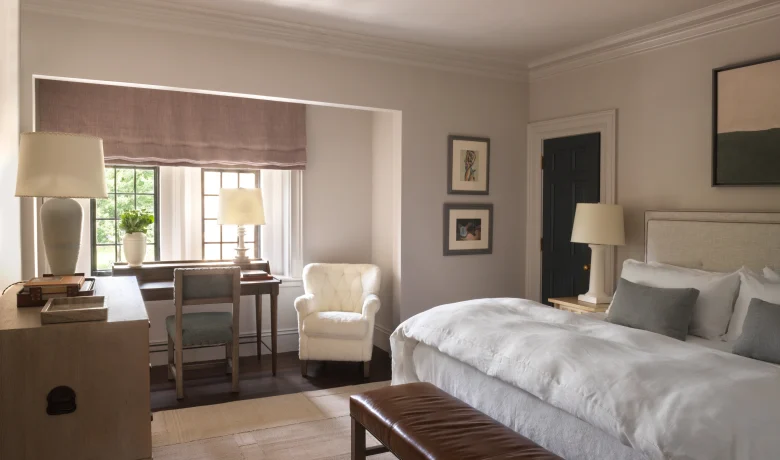 Neutral-toned hotel bedroom with two white beds, a brown leather bench at the foot, and cream armchairs by a desk near a window with a lavender shade; soft lighting and framed art on the walls.
