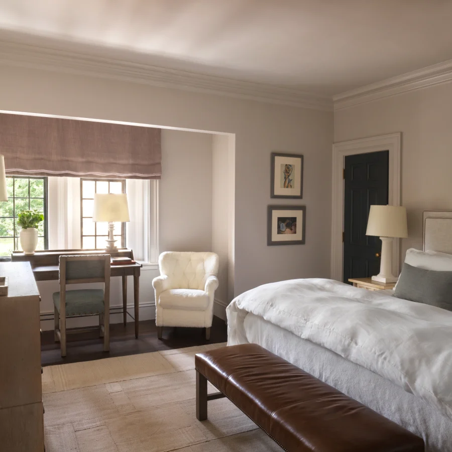 Neutral-toned hotel bedroom with two white beds, a brown leather bench at the foot, and cream armchairs by a desk near a window with a lavender shade; soft lighting and framed art on the walls.