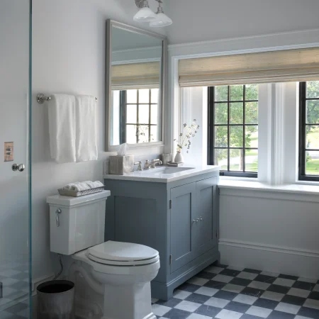 Modern bathroom with a blue-grey vanity, marble countertop, and a glass shower enclosure; large window with shades and a black-and-white checkered floor.