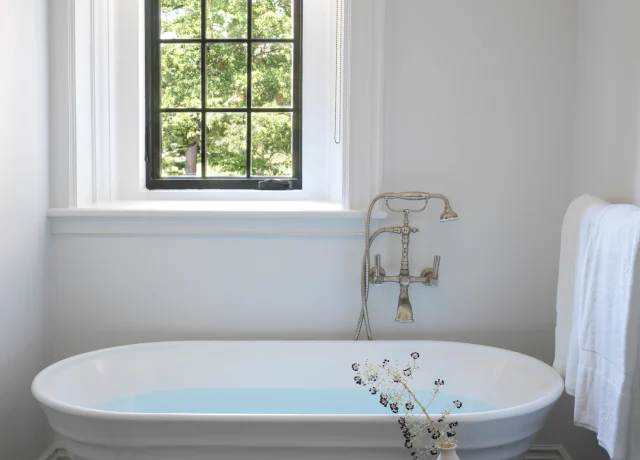 White freestanding bathtub in a bright bathroom with a black-framed window, woven shade, and a dark pedestal table with a vase and candle.