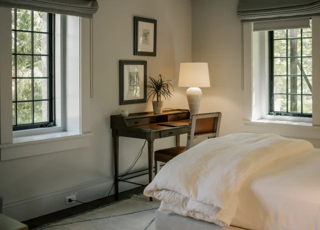 Corner of a bedroom with a wooden desk, lamp, potted plant, and two framed pictures on a gray wall, by a window.