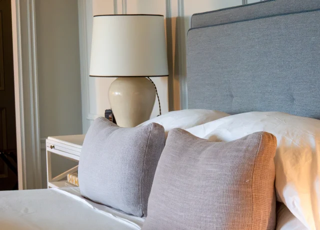 Bed with white duvet and two light gray throw pillows in front of a gray upholstered headboard and paneled wall, bedside lamp on a white nightstand to the left.