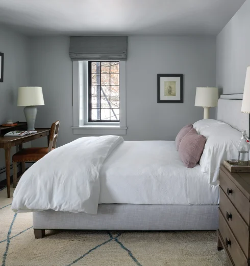 Cozy gray bedroom with a white duvet, mauve pillows, and two dark wood nightstands—lamps on each side and a window at the far wall.