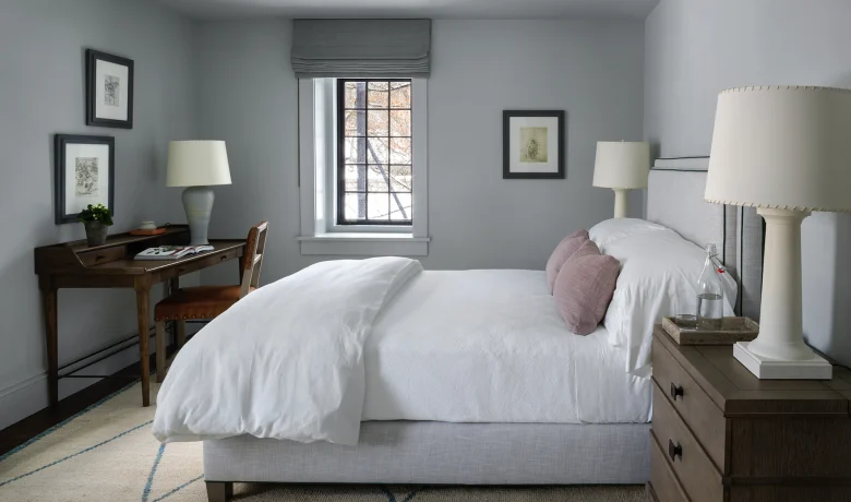 Cozy gray bedroom with a white duvet, mauve pillows, and two dark wood nightstands—lamps on each side and a window at the far wall.