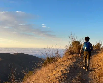 Hiker with backpack standing on a rocky hillside at sunset, overlooking a coastline with bare shrubs.