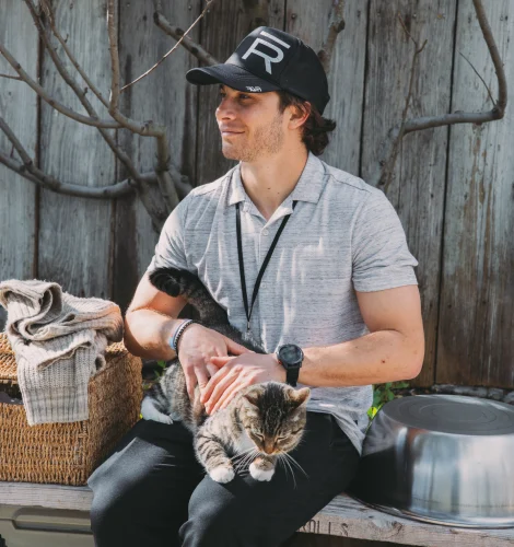 Man wearing a gray polo and black cap sitting outdoors, gently holding a tabby cat on his lap beside woven baskets and a wooden fence backdrop.
