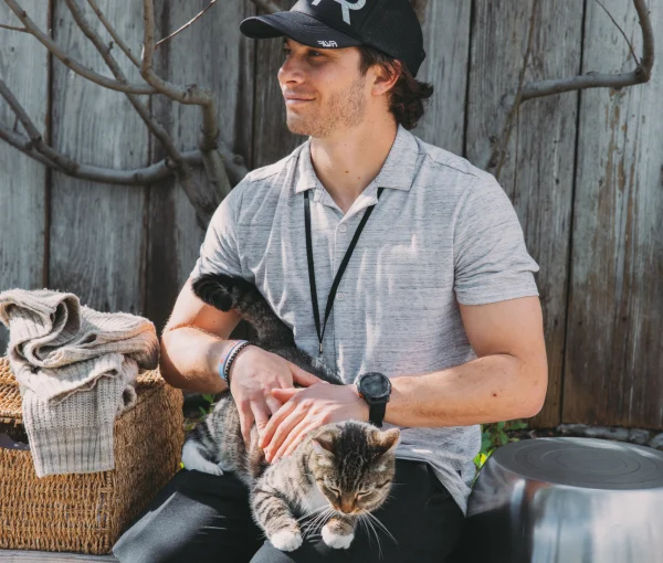Man wearing a gray polo and black cap sitting outdoors, gently holding a tabby cat on his lap beside woven baskets and a wooden fence backdrop.