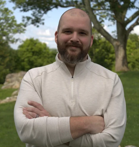 Man with a beard and shaved head, wearing a beige fleece, arms crossed in a park.