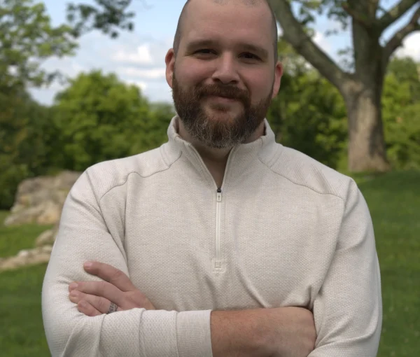 Man with a beard and shaved head, wearing a beige fleece, arms crossed in a park.