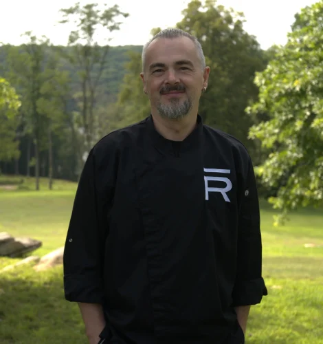 Man with gray beard and mustache wearing a black chef coat, standing in a sunlit park with trees in the background.