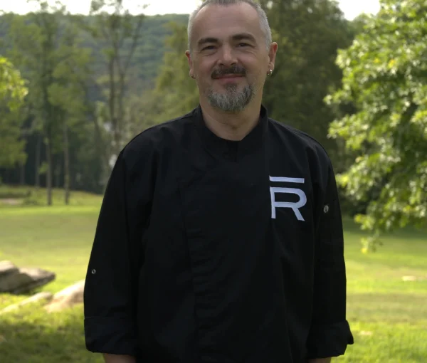 Man with gray beard and mustache wearing a black chef coat, standing in a sunlit park with trees in the background.