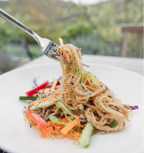 Pile of glass noodles with julienned carrots, cucumbers, and peppers on a white plate outdoors.