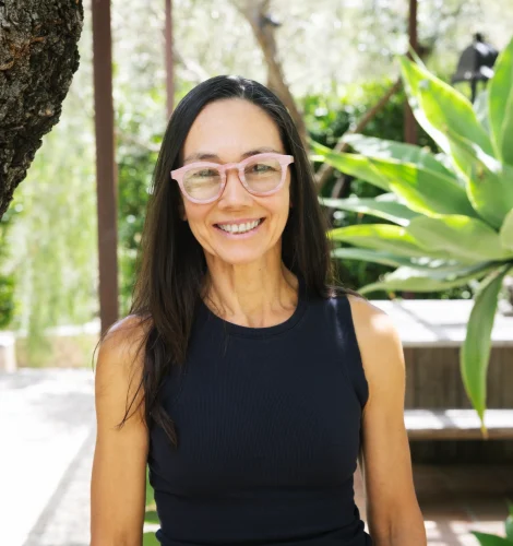 Smiling woman with pink glasses standing outdoors in a sunny garden with greenery and a tree nearby.