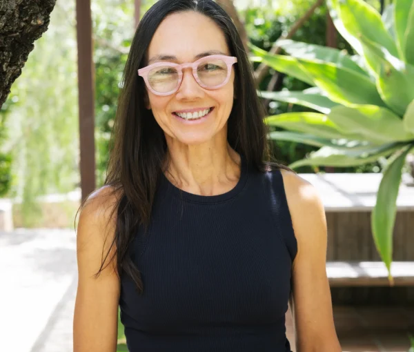 Smiling woman with pink glasses standing outdoors in a sunny garden with greenery and a tree nearby.