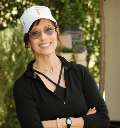 Smiling woman standing outdoors with arms crossed, wearing a white cap, sunglasses, and a black zip-up top.