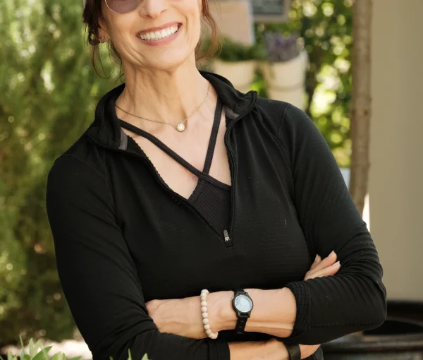 Smiling woman standing outdoors with arms crossed, wearing a white cap, sunglasses, and a black zip-up top.