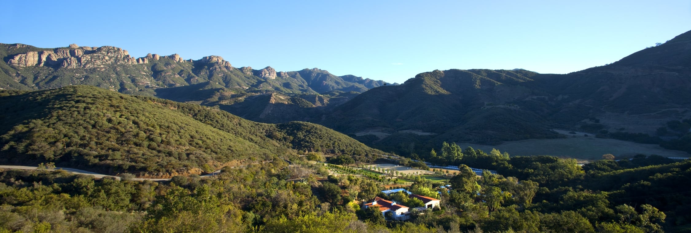 Wide view of rolling green hills and distant mountains under a clear blue sky, with a small cluster of buildings at the valley bottom.