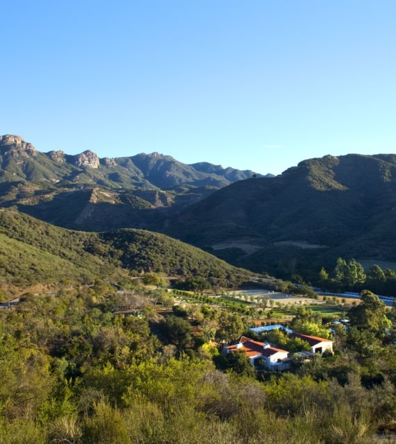 Wide view of rolling green hills and distant mountains under a clear blue sky, with a small cluster of buildings at the valley bottom.