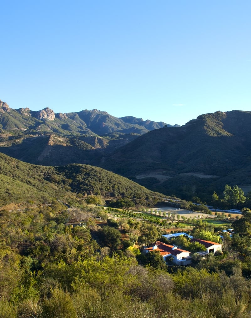 Wide view of rolling green hills and distant mountains under a clear blue sky, with a small cluster of buildings at the valley bottom.