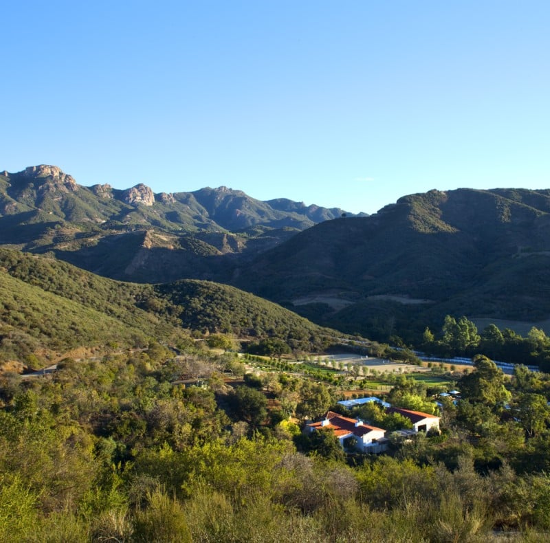Wide view of rolling green hills and distant mountains under a clear blue sky, with a small cluster of buildings at the valley bottom.