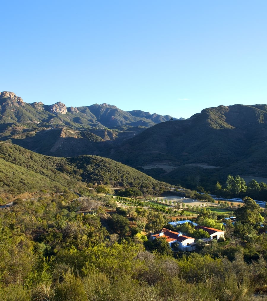 Wide view of rolling green hills and distant mountains under a clear blue sky, with a small cluster of buildings at the valley bottom.