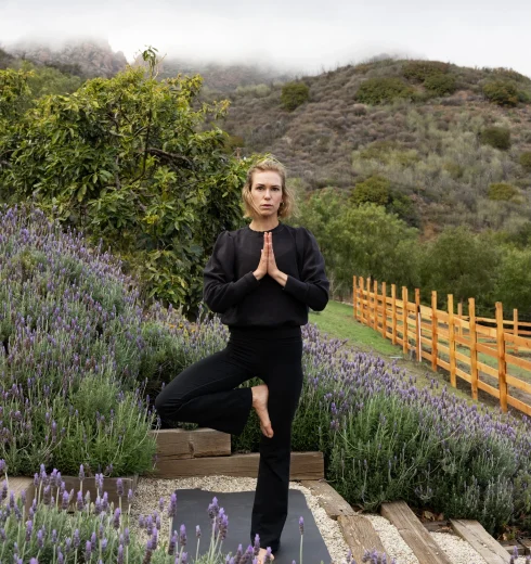 Woman in black yoga clothes practices tree pose among lavender plants with hills and cloudy sky behind her, outdoors.