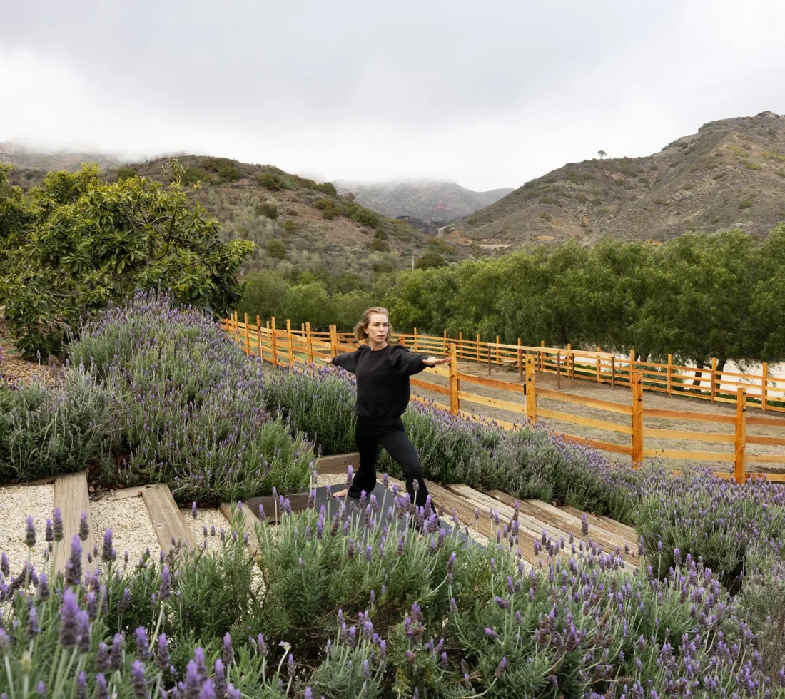 Woman in a black top stands in a lavender garden with purple flowers and a wooden fence, hills in the background under a cloudy sky.