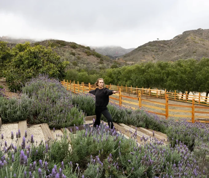 Woman in a black top stands in a lavender garden with purple flowers and a wooden fence, hills in the background under a cloudy sky.