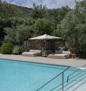 Poolside lounge area with white umbrella and cushioned wooden sofas, stone wall behind, and olive trees under a blue sky.