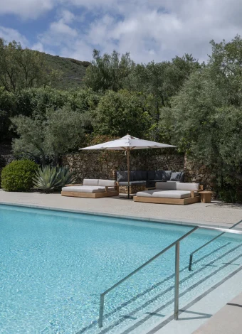 Poolside lounge area with white umbrella and cushioned wooden sofas, stone wall behind, and olive trees under a blue sky.
