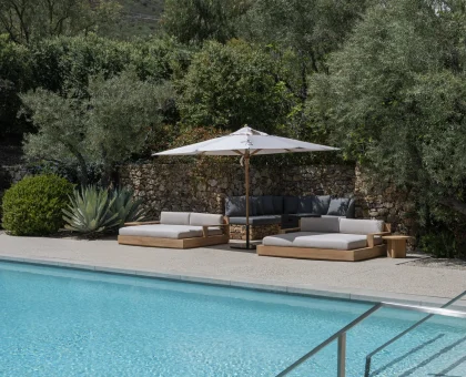 Poolside lounge area with white umbrella and cushioned wooden sofas, stone wall behind, and olive trees under a blue sky.