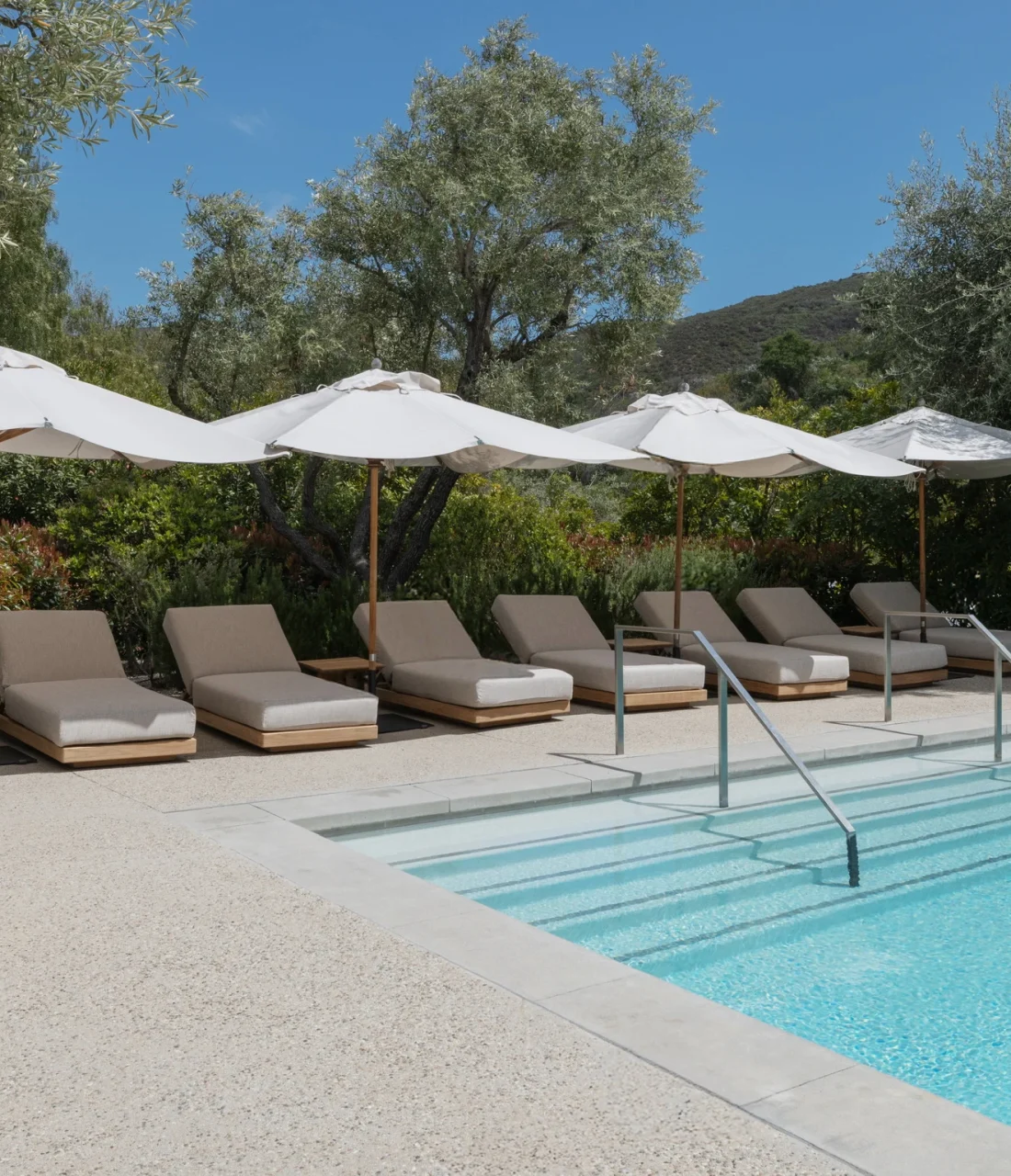 Row of beige pool loungers under white umbrellas by a blue pool with trees and hills in the background.