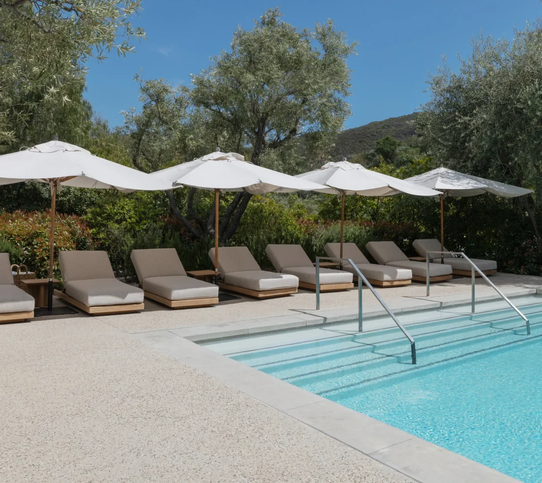 Row of beige pool loungers under white umbrellas by a blue pool with trees and hills in the background.