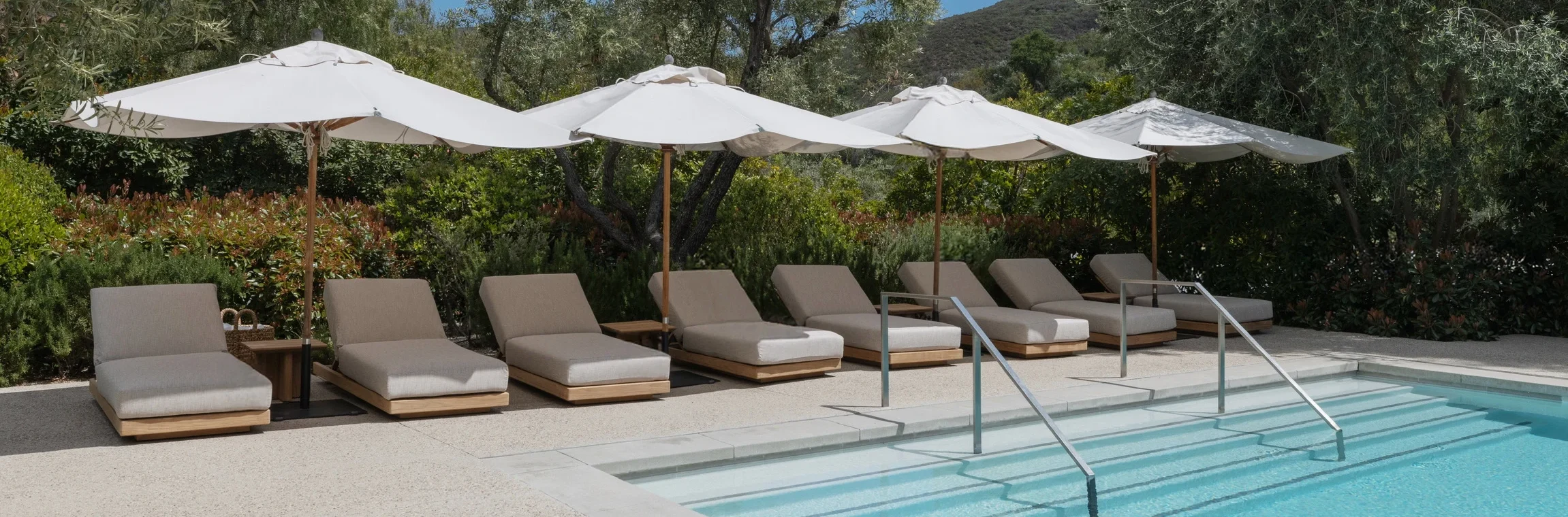 Row of beige pool loungers under white umbrellas by a blue pool with trees and hills in the background.
