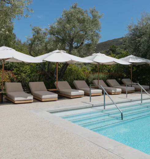 Row of beige pool loungers under white umbrellas by a blue pool with trees and hills in the background.